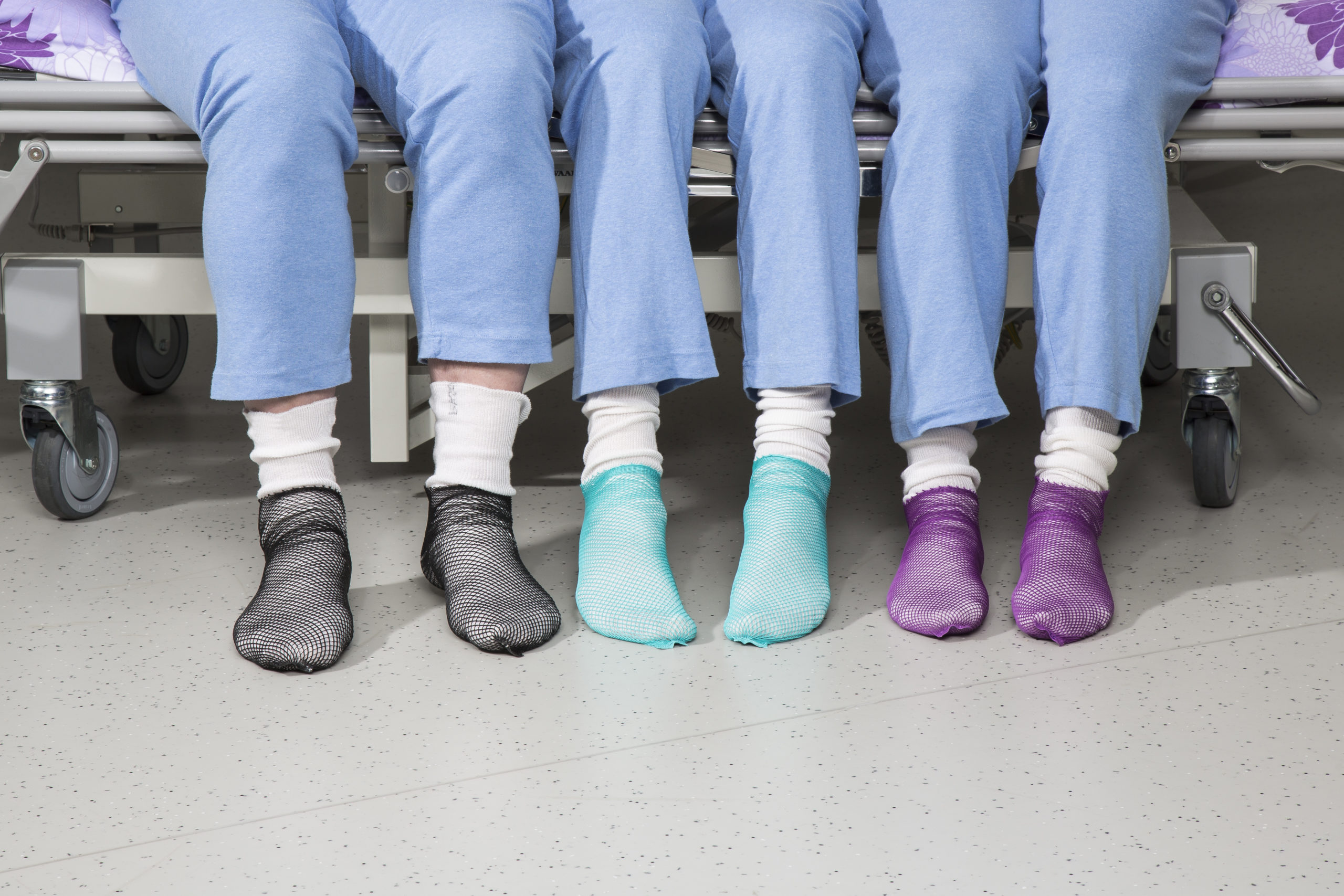 Sinihousuiset henkilöt istuvat vierekkäin sairaalasängyllä eri väriset Pitosukat jalassaan